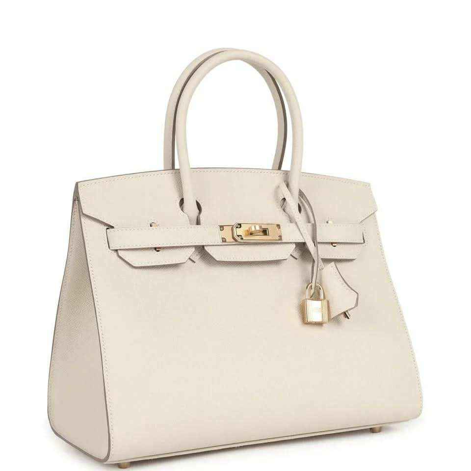 Top-handle leather handbag with gold hardware, front flap and lock, light neutral.