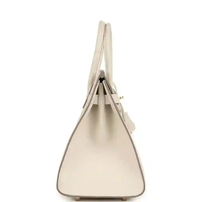 Top-handle leather handbag with gold-toned hardware, tapered shape, light neutral.