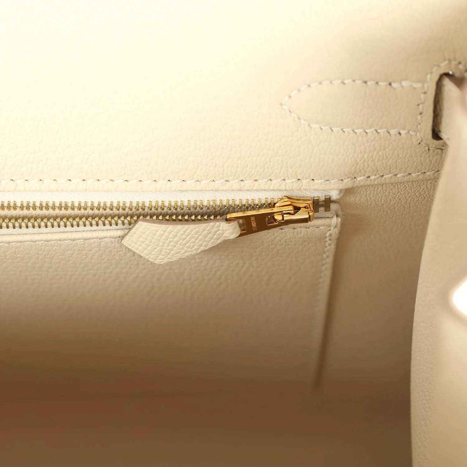 Leather handbag zipper with gold-toned pull and stitching, light neutral.