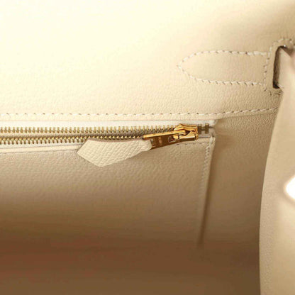 Leather handbag zipper with gold-toned pull and stitching, light neutral.
