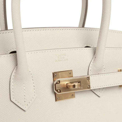 Leather handbag front clasp with gold-toned hardware and stitching, light neutral.