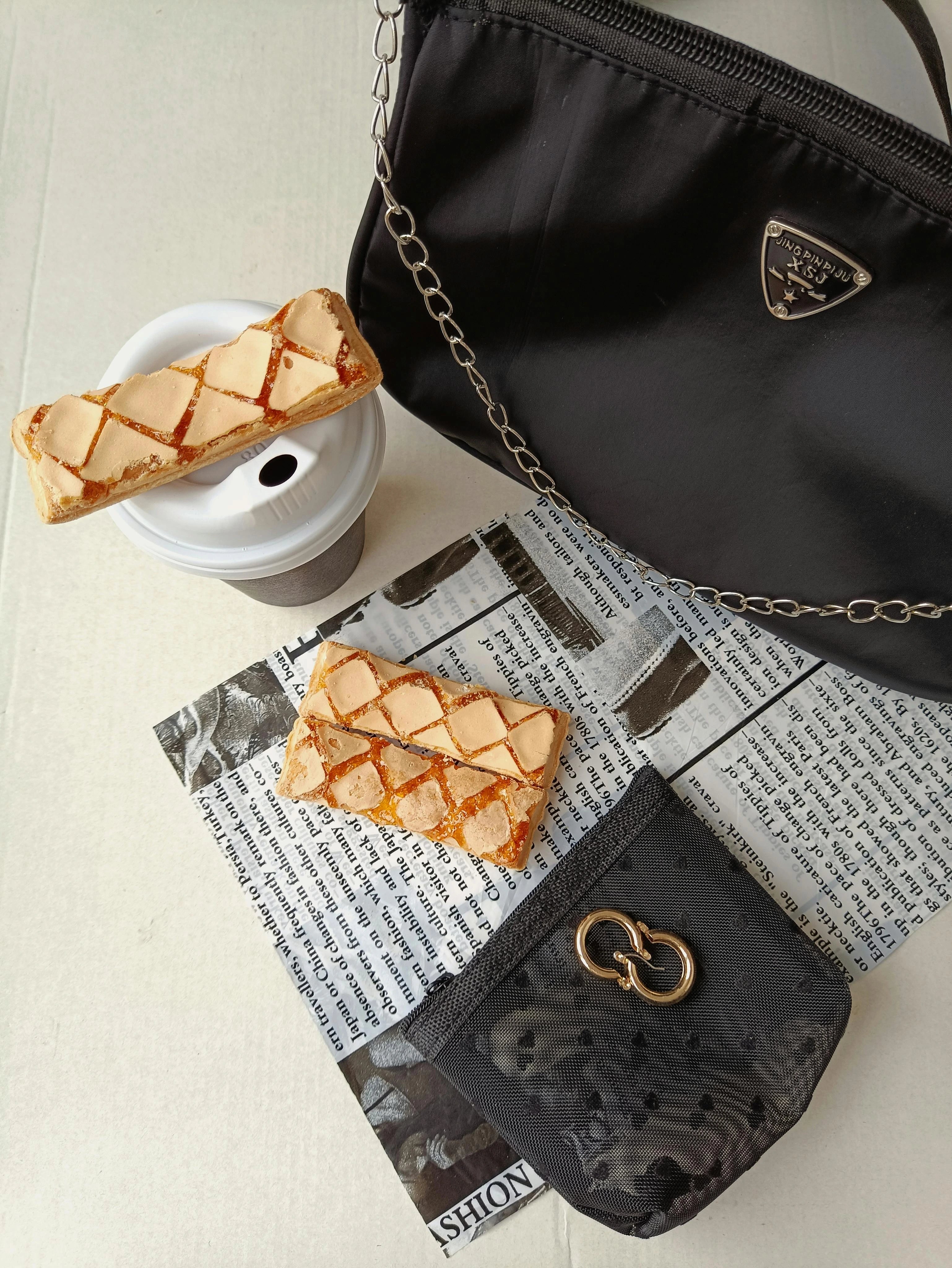 Black handbag with a chain detail, a small pouch, and pastries on a newspaper surface.